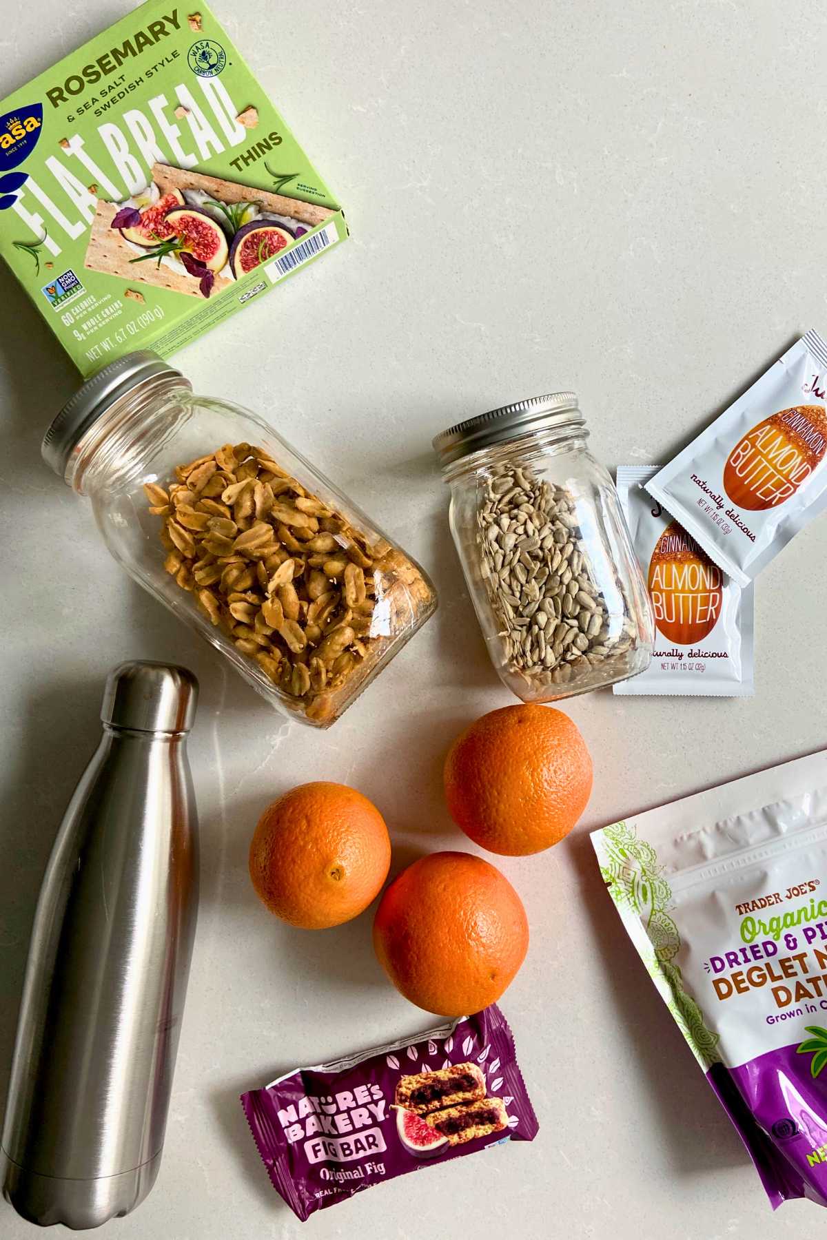 Water bottle, jars of nuts, oranges, nut butter packets, dates and crackers on a counter