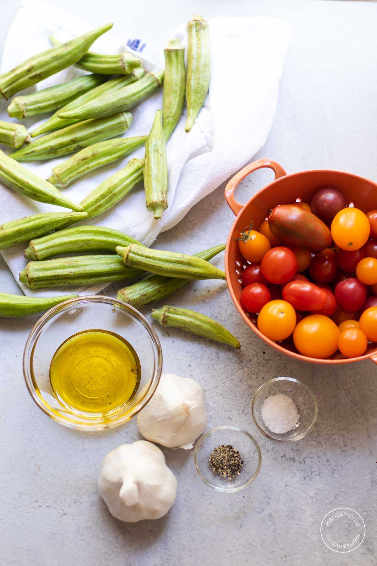 Ingredients for the recipe on a gray backdrop including okra, tomatoes, oil, salt, pepper, and garlic.