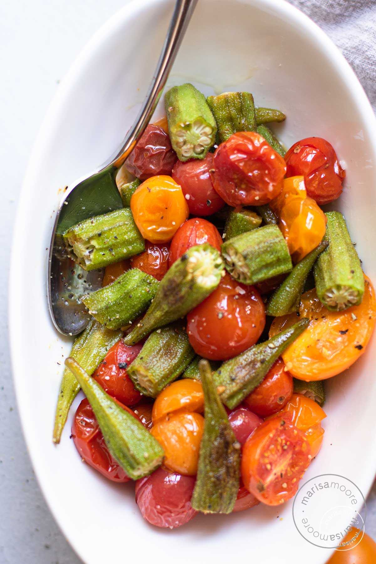Roasted okra and tomatoes in a white bowl with a spoon.