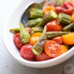 Roasted okra and tomatoes in a white oval bowl with a serving spoon.