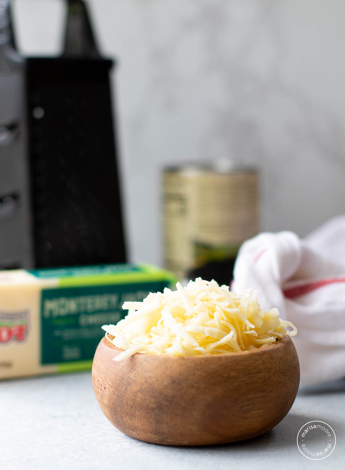Wooden bowl filled with shredded cheese and a block of cheese and a grater in the background