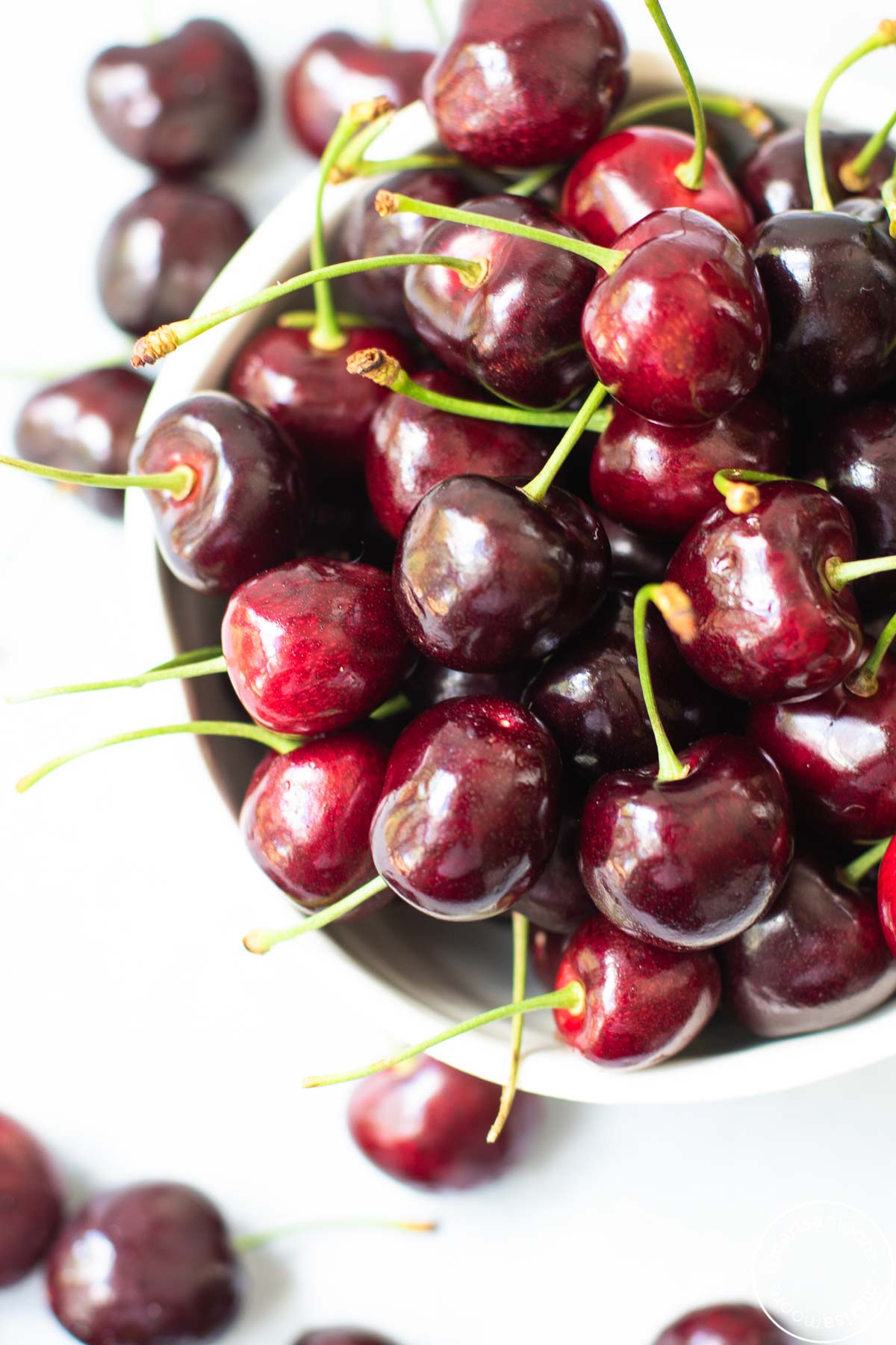 fresh dark sweet cherries with green stems in a white bowl with cherries scattered around 