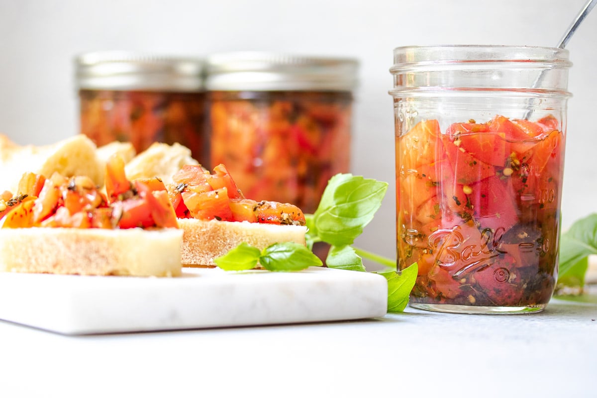 Bruschetta tomatoes in a jar with a snack board and pieces of crostini topped with marinated tomatoes and basil