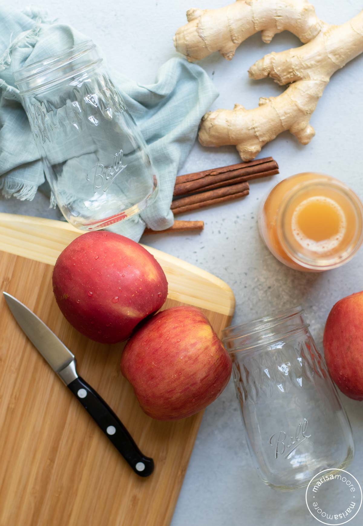 two red apples on a cutting board with a knife, 2 fluted jar and the apple juice and cinnamon ingredients 