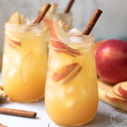 two fluted jars with the apple mocktail inside garnished with cinnamon sticks and apple slices