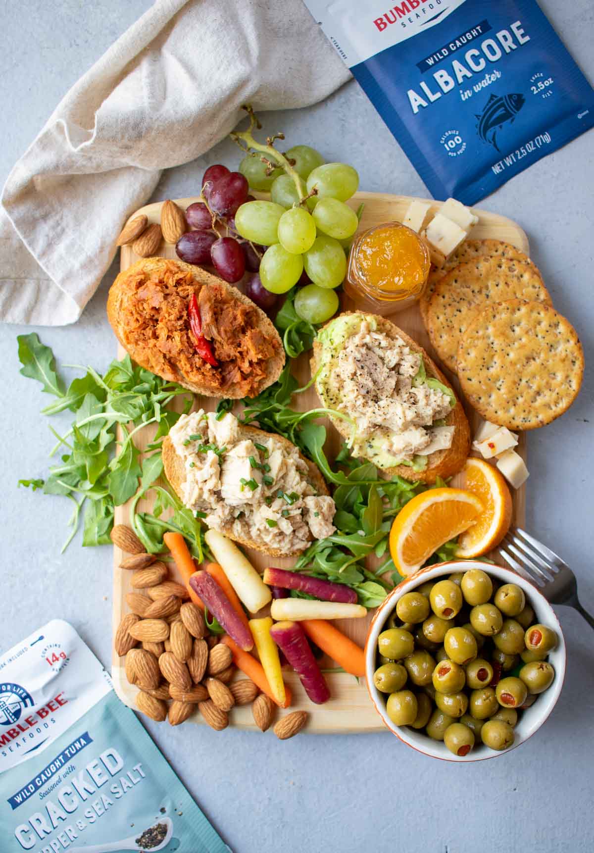 Snack board with tuna toasts, fruit, vegetables, nuts and packets of Bumble Bee Tuna