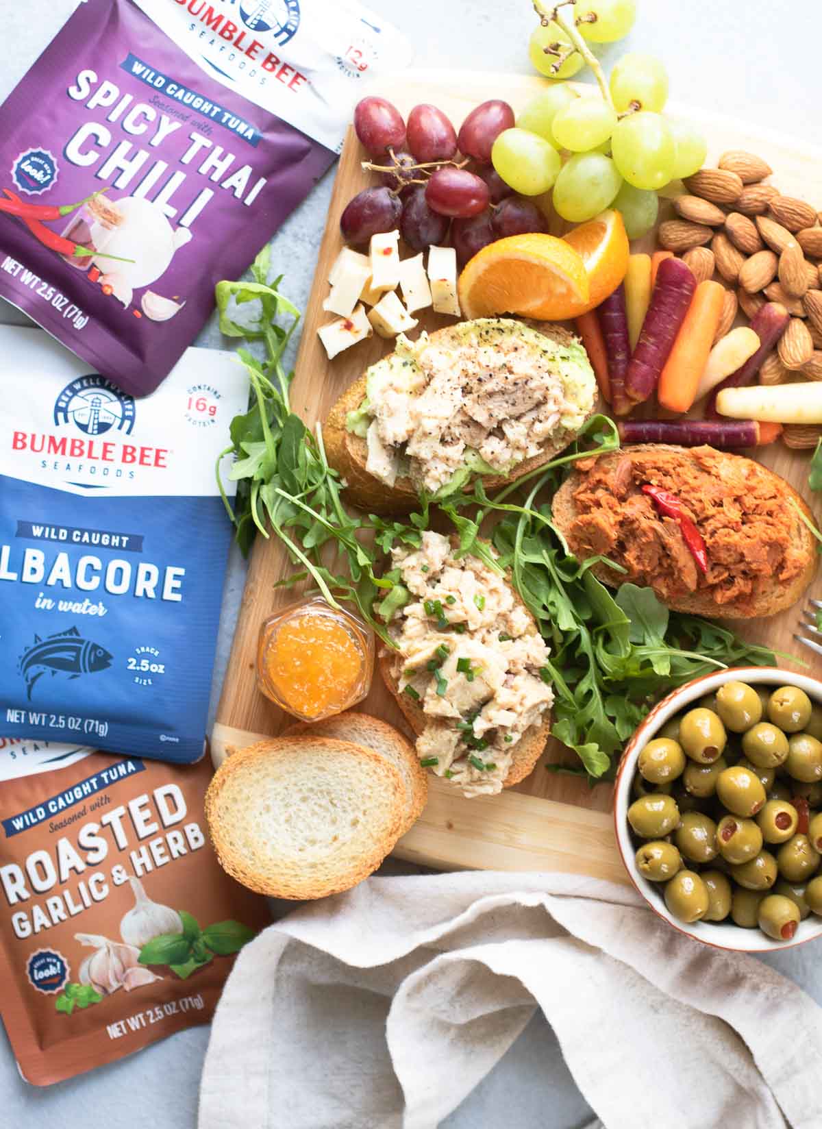 Crowded board with all tuna toasts and ingredients with packets of Bumble Bee Tuna on the side