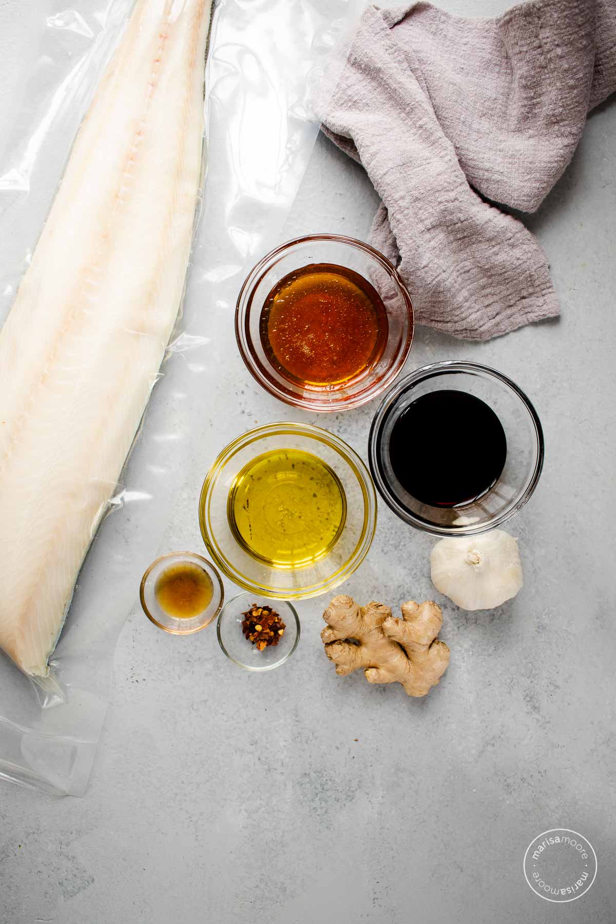 Ingredients on a grey board. Sablefish in plastic, honey, oil, soy sauce, sesame oil, red pepper flakes in bowls, whole pieces of garlic and ginger.