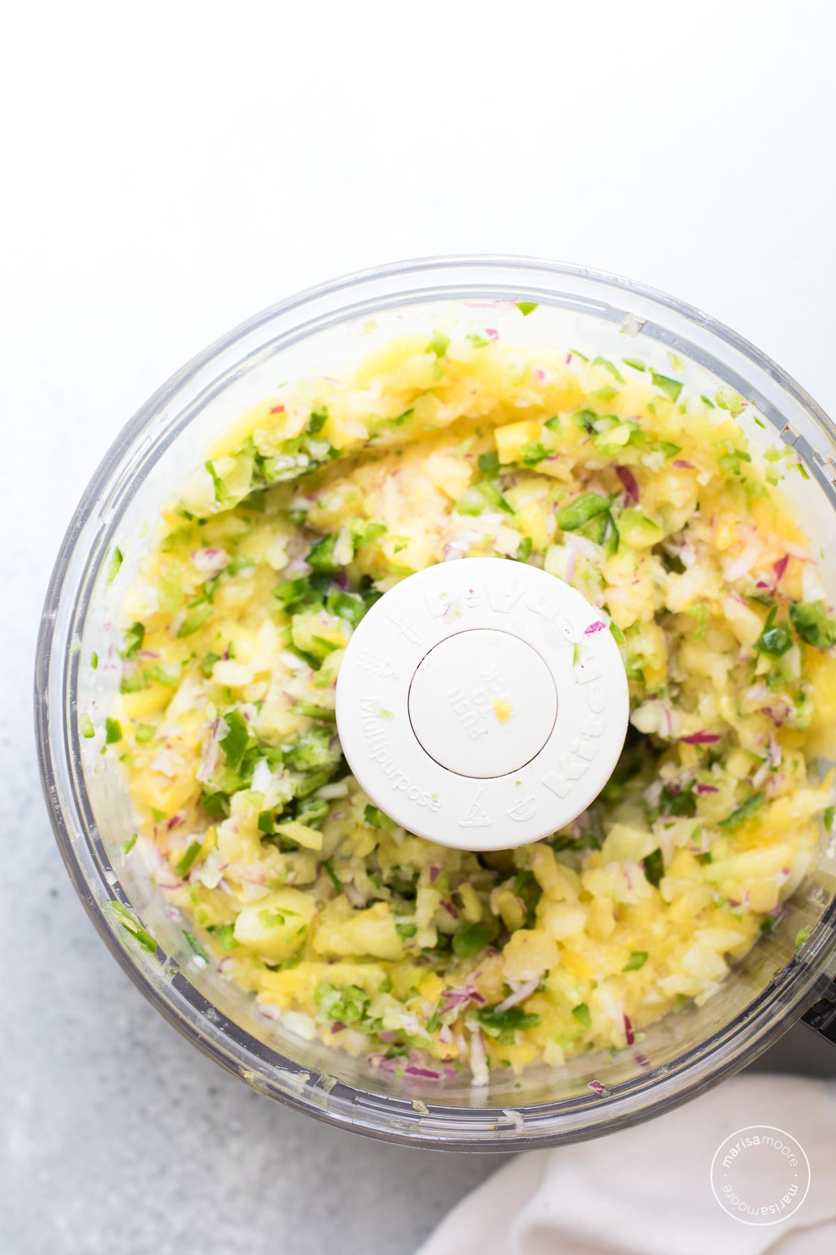 Bowl of a food processor with chopped pineapple, onion and jalapeno