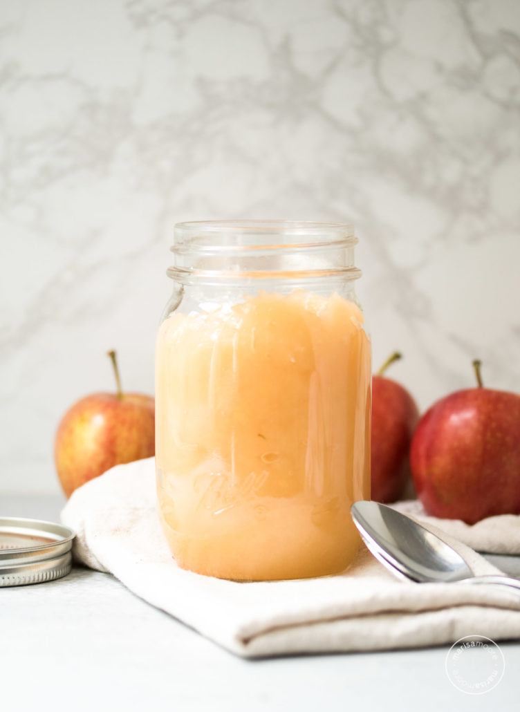 Open jar of applesauce with a spoon and apples in background.