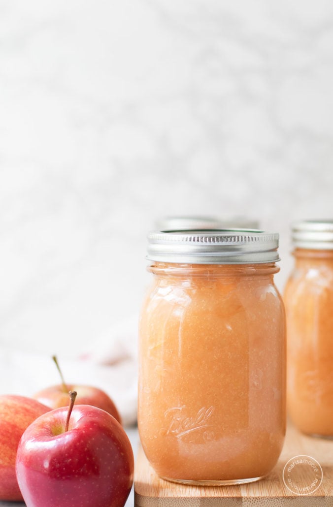 Applesauce in 3 pint jars on a cutting board with red apples