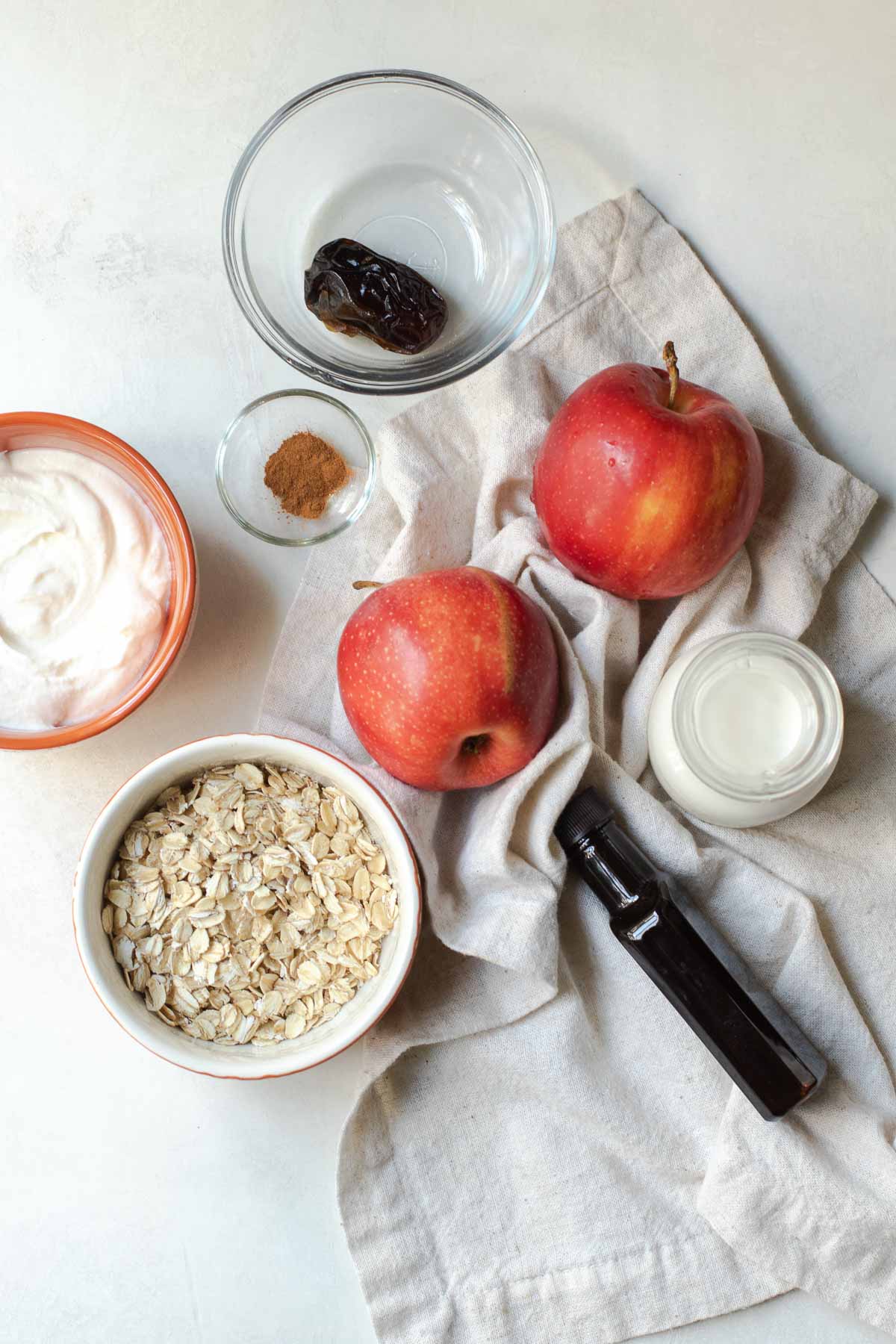 All ingredients on a gray background: 1 date, two apples, cinnamon, milk, vanilla extract bottle, oats, and plain yogurt.