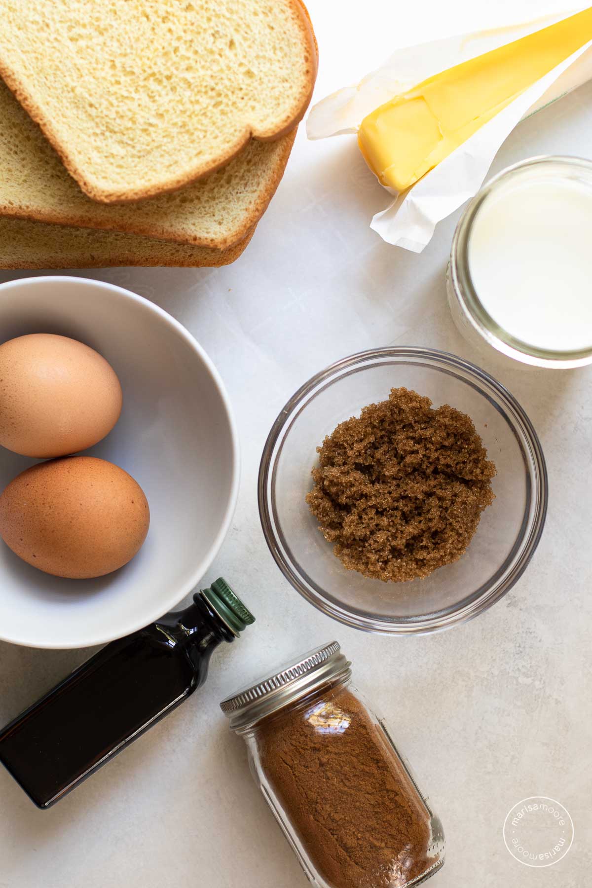 Ingredients for the baked French toast including bread, butter, milk, brown sugar, cinnamon, vanilla in a bottle and eggs in a bowl.