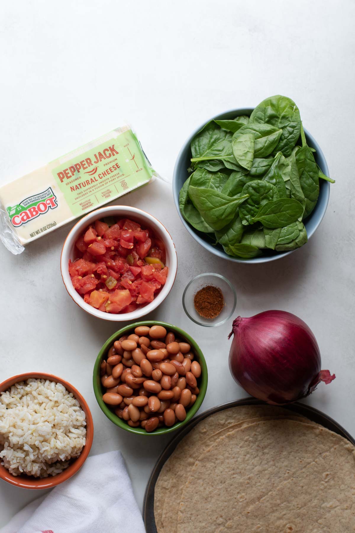 Bar of Cabot Pepper Jack Cheese and burrito ingredients in bowls with spinach, tomatoes, pinto beans, rice and chili powder, tortillas and a whole onion