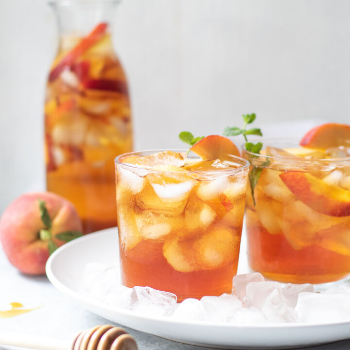 Peach iced tea in glasses surrounded by ice with a carafe of peach iced tea in the background.