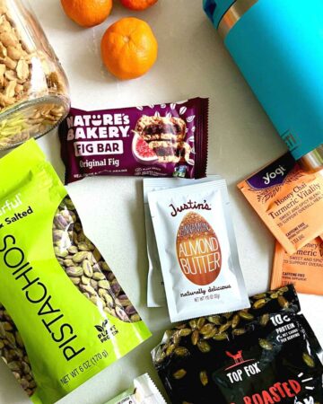 Flatlay of jar of peanuts, tangerines, tea packets, water bottle, nut butter packets, bags of pumpkin seeds and pistachios