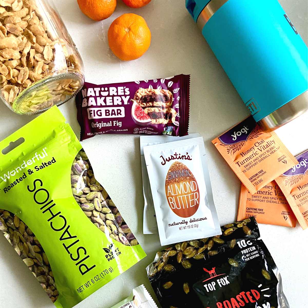 Flatlay of jar of peanuts, tangerines, tea packets, water bottle, nut butter packets, bags of pumpkin seeds and pistachios