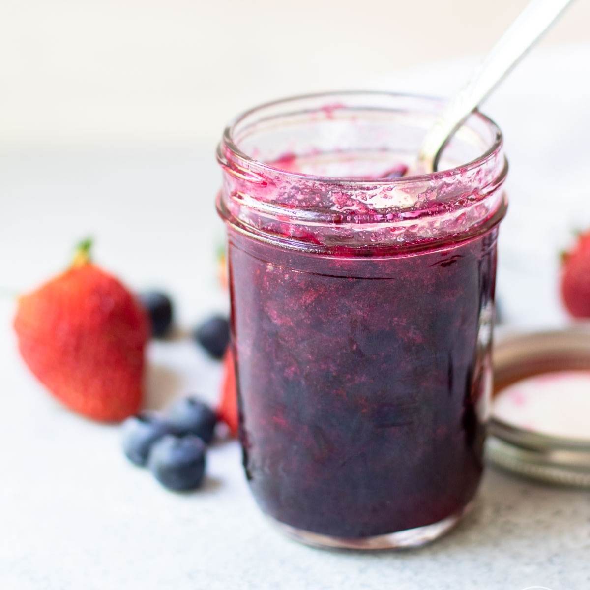 Jam in a pint-sized jar with a spoon inside and scattered strawberries and blueberries surrounding it.