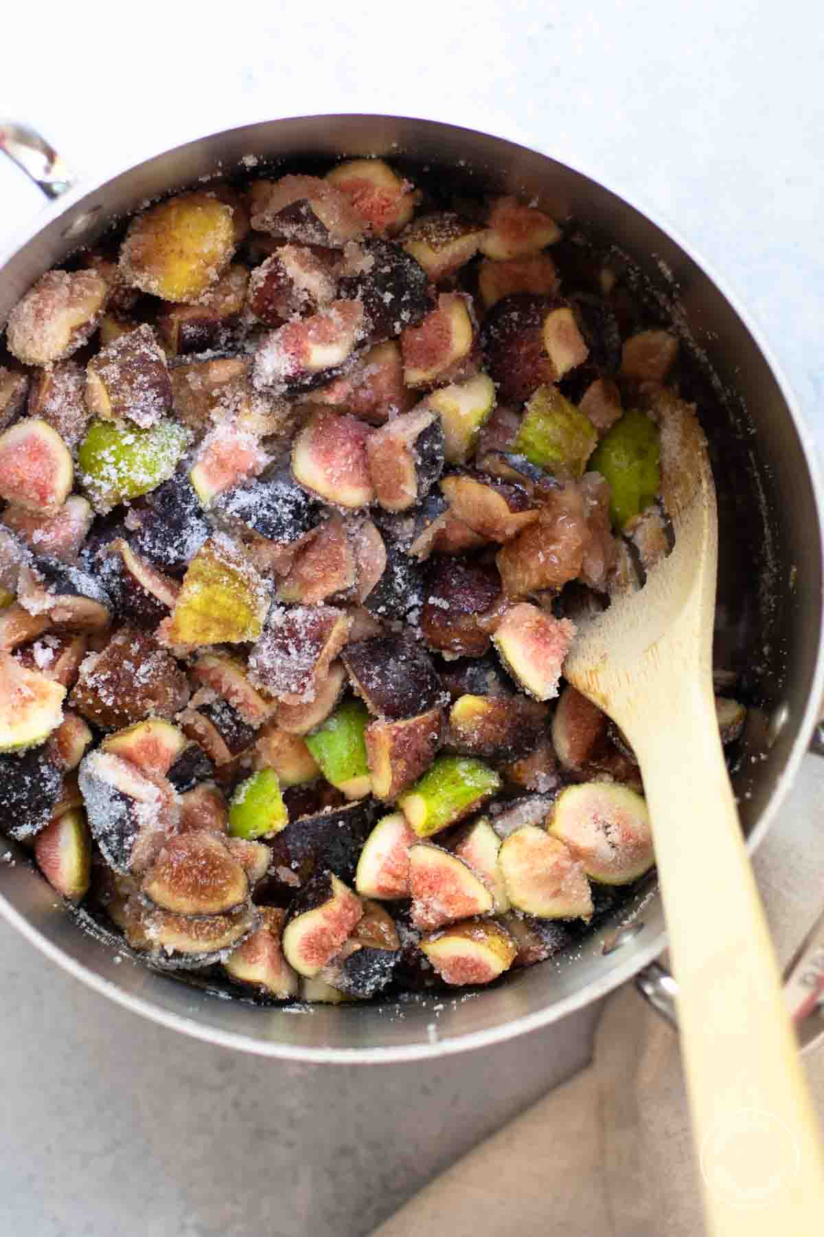 Cut figs of many colors with sugar sprinkled on top in a stainless steel pot with a wooden spoon.