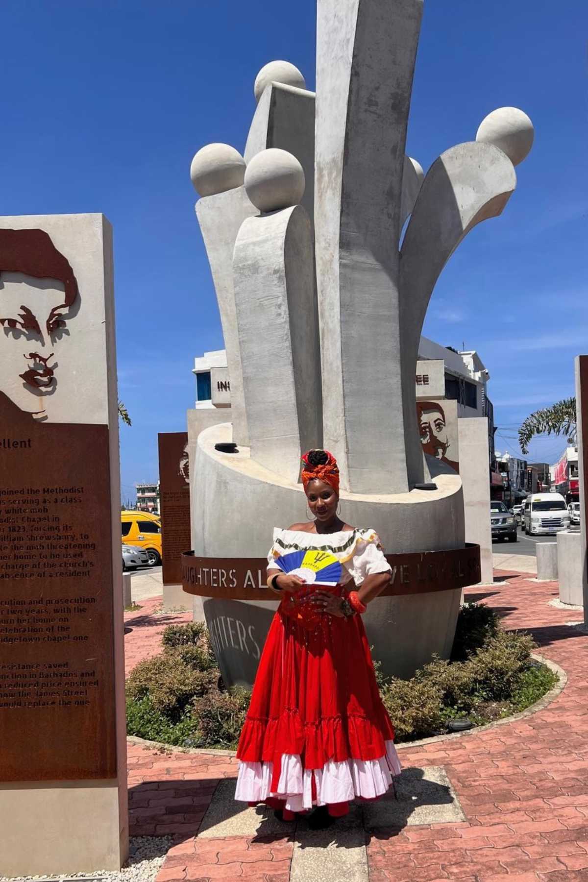 Bajan woman tour guide dressed in traditional attire of a red flow skirt, white blouse and red head wrap. She is standing in Heroes Square.  